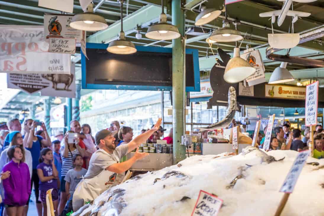 Photo of a busy fish market with a man wearing an apron throwing a large fish across the room. Pike Place is a must during your 48 hours in Seattle.