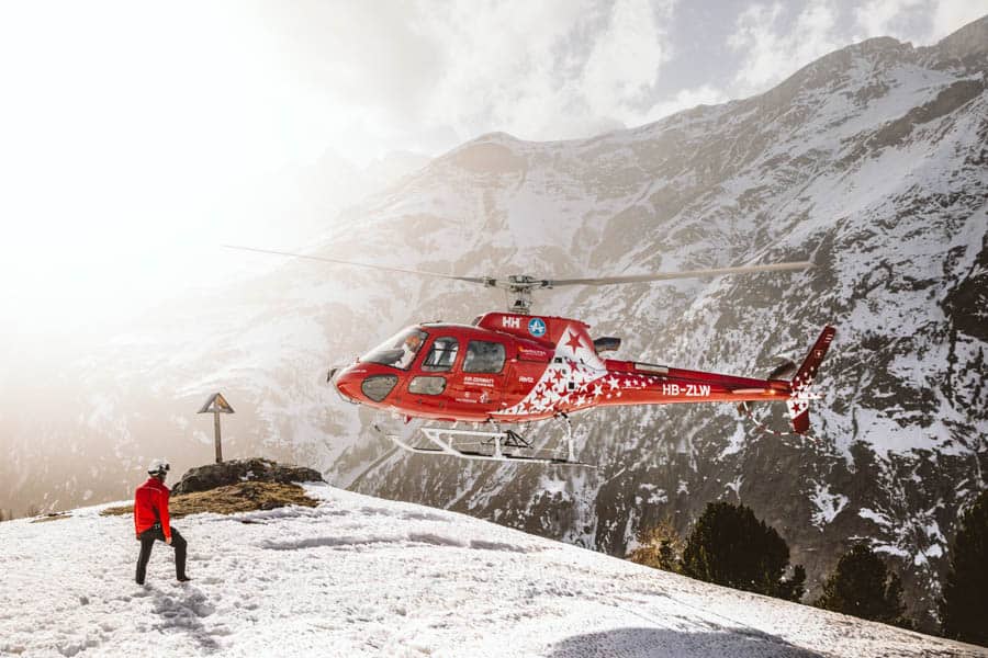Photo of a man on a snowy mountain walking towards a red helicopter about to land, with a snow-capped mountain in the background.