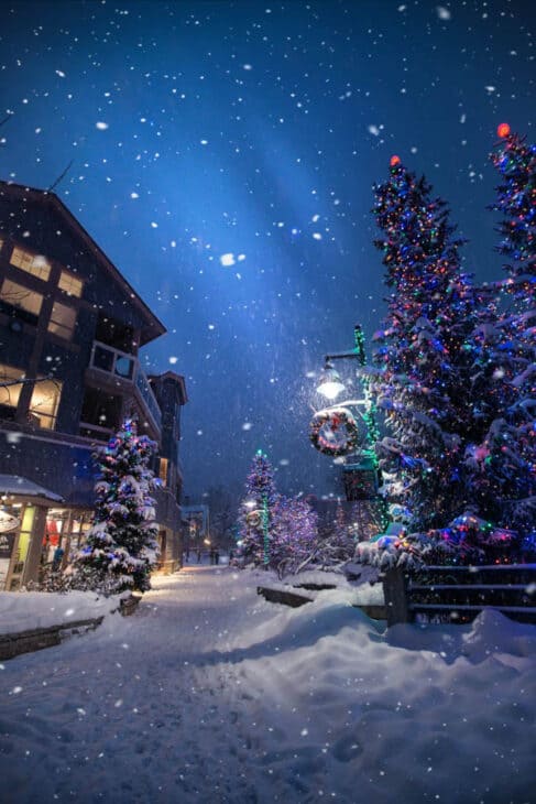 Photo of a town at night with snow falling and christmas lights on the trees.
