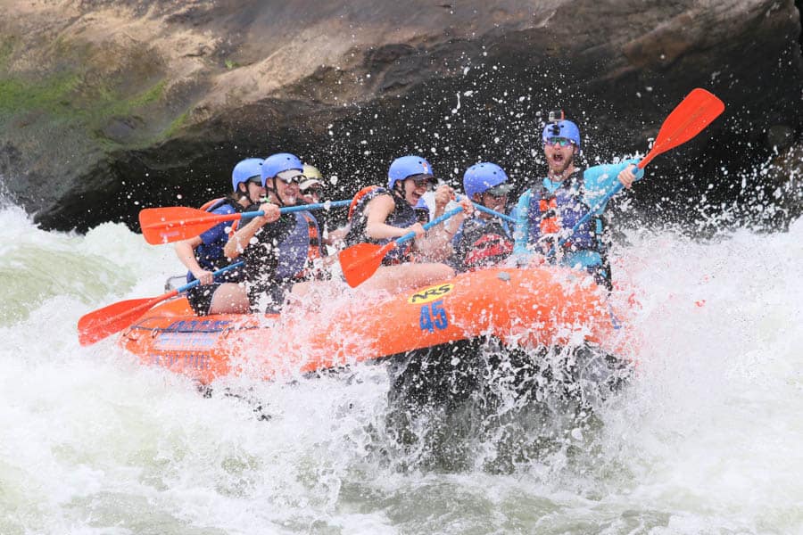 Photo of a group of people on an inflatable boat with paddles drifting down a river with rapids and water splashing. 