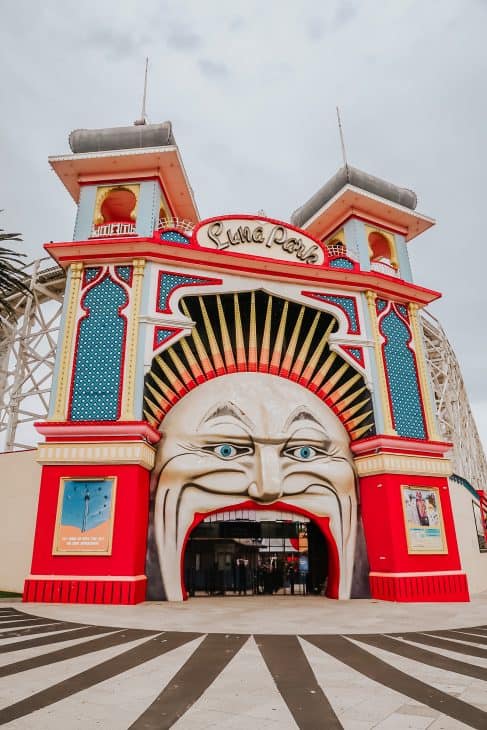 Photo of the facade of Luna Park in Melbourne Australia. The facade has the face of a clown with red lips and blue eyes and a crown of spikes in red, yellow and orange. The pillars either side are adorned with blues, reds and yellow. Luna Park in St. Kilda is an iconic landmark that makes for an awesome Instagram spot.