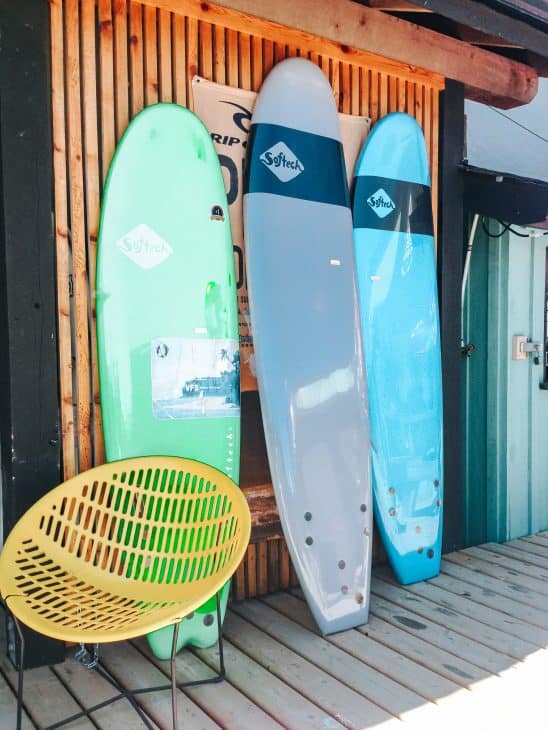 Photo of three surfboards in green grey and blue leaning against a wooden wall with a yellow beach chair in fron of them. Surfing is one of the most popular activities in Tofino.
