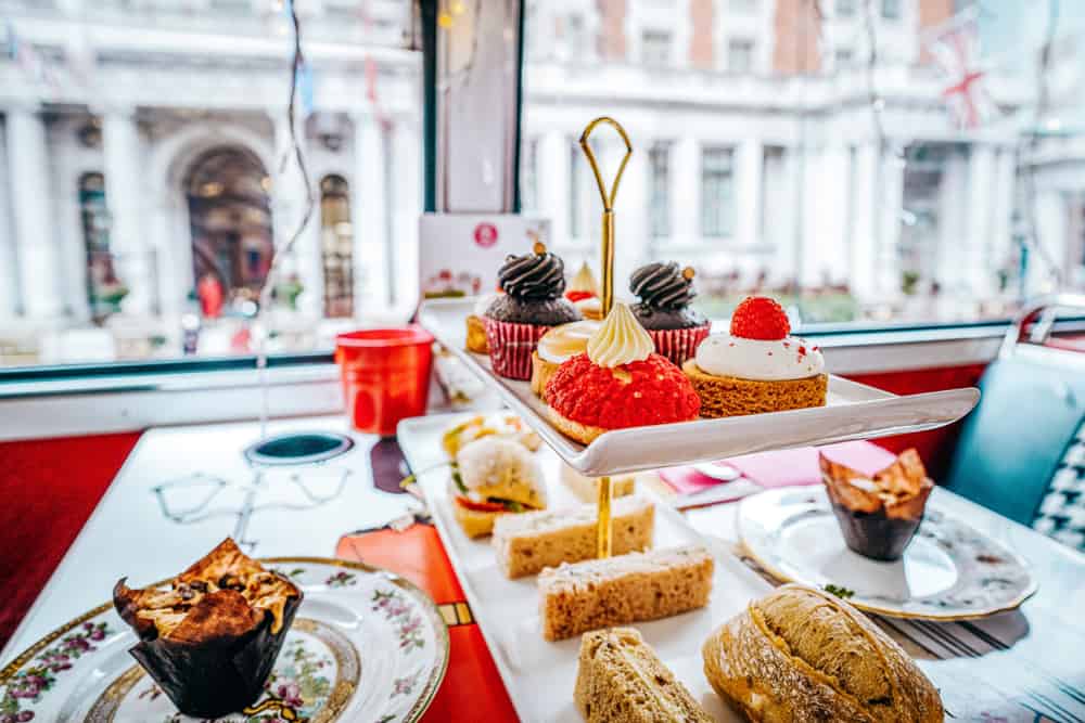 Photo of a two tiered tray filled with cakes and sandwiches. On the top tier are two chocolate cupcakes and two pastries, one with red icing and one with white icing a raspberry on top. On the bottom tier are finger sandwiches and bread buns. Either side of the tray are two ornate plates with muffins on. 