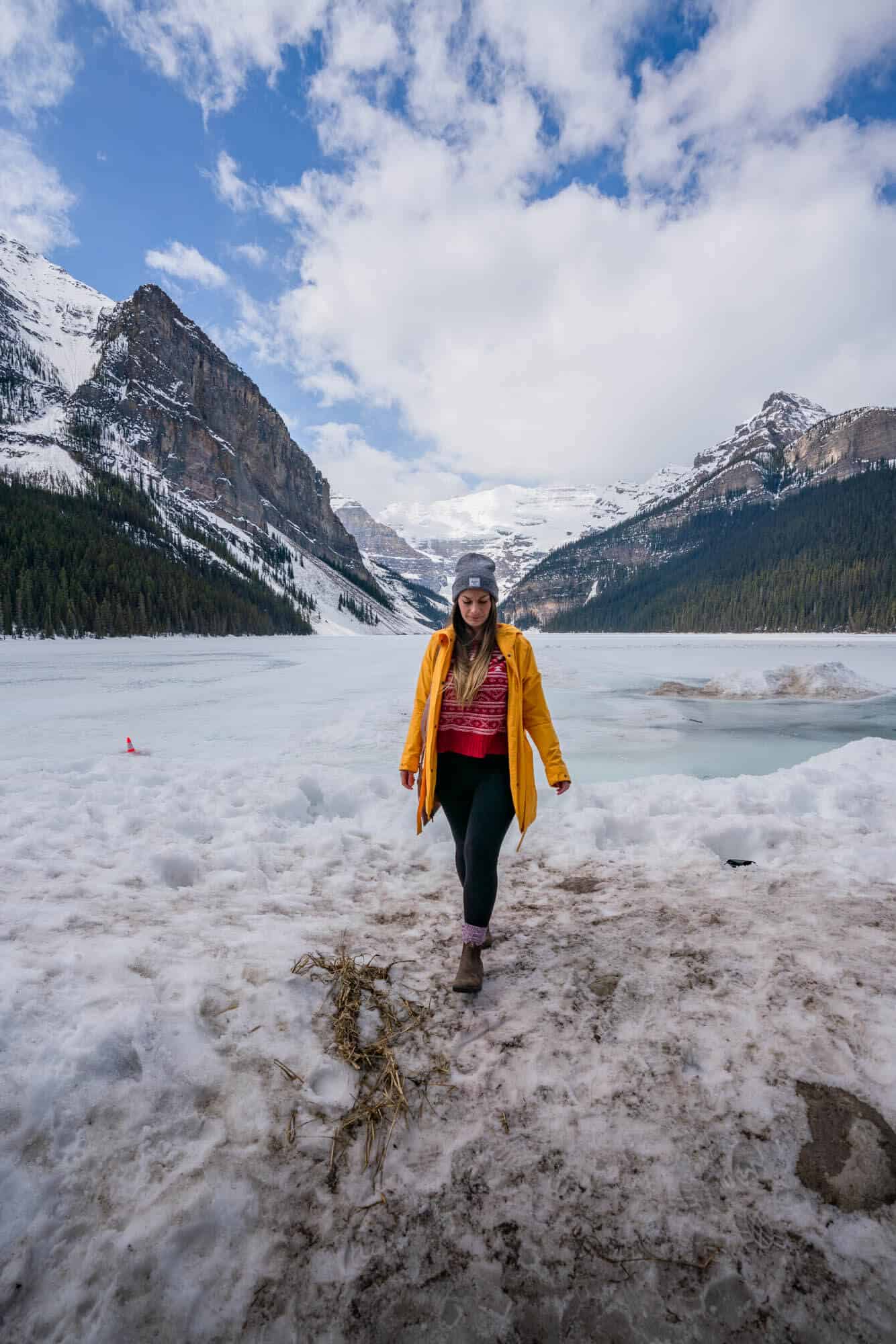 Photo of a woman walking towards the camera on a snowy and frozen lake with. aglacier and two mountain peaks in the background.
