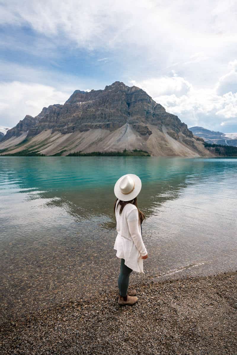 A woman wearing a white hat and white cardigan standing at the shore of a blue glacial lake while looking out towards a mountain peak. 