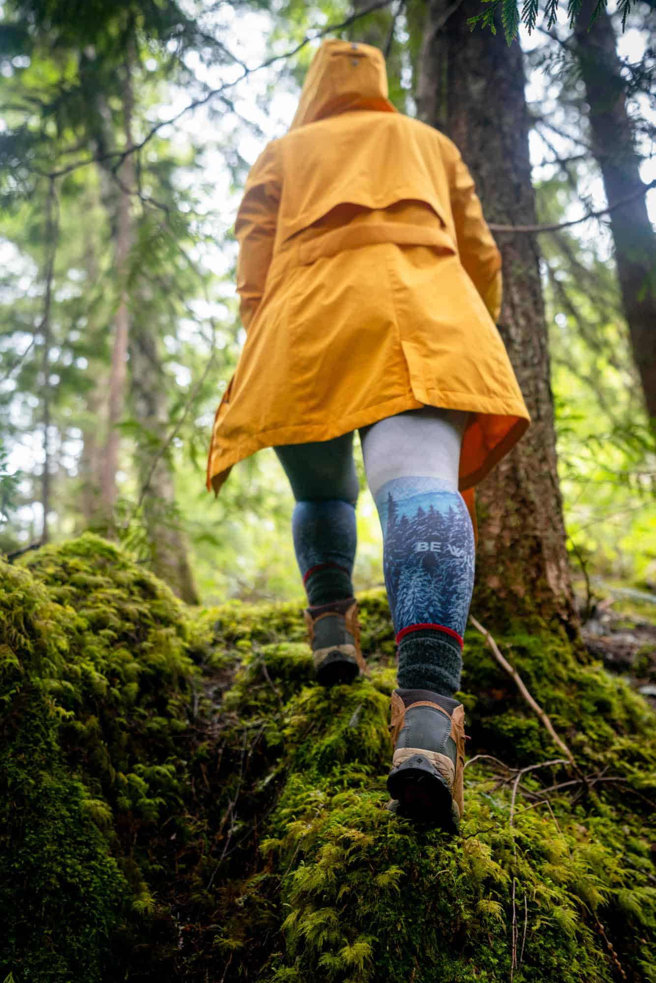 Photo taken from below of a woman wearing a yellow rain coat walking in a mossy green forest. 