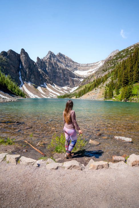 Photo of a woman standing at a lake shore of a green coloured lake with rocky mountain peaks in the background. 