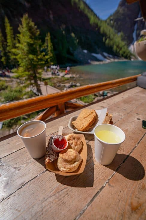 Photo of a wooden table with 2 drinks, a sandwich and two scones with a view of the lake in the background.