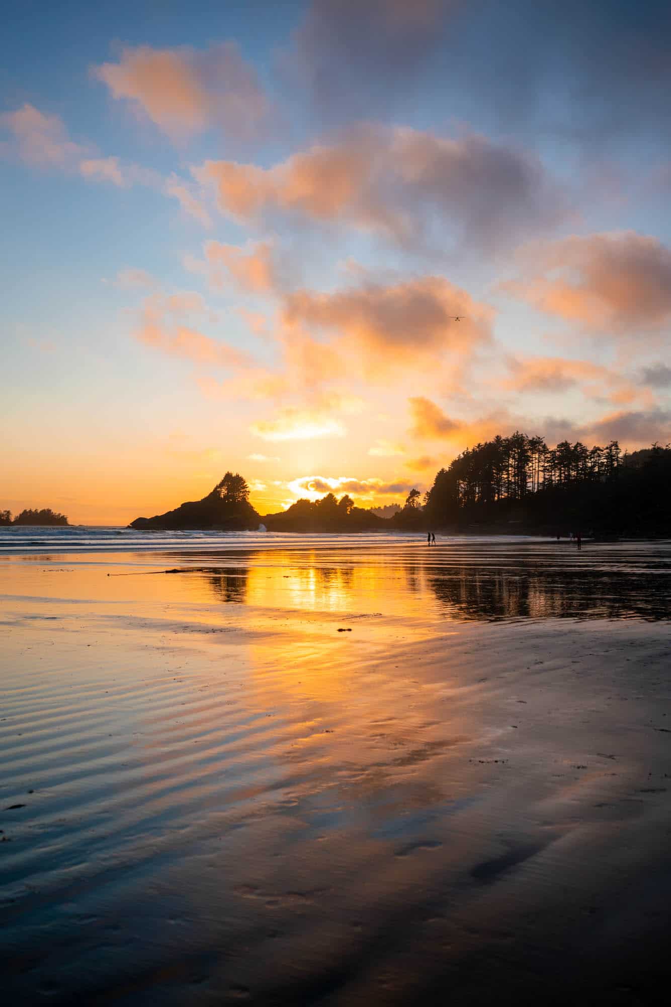 Yellow, orange and pink hues fill the sky at sunset on Cox Bay Beach with the ocean and wet sand reflecting the sunset colour. Cox Bay is a must visit when ending your Vancouver Island road trip in Tofino. 