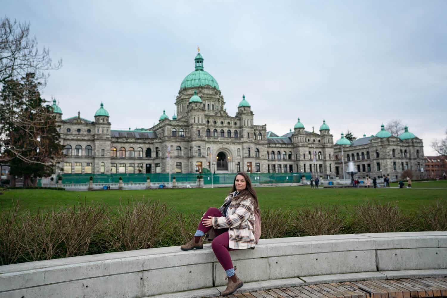 Woman sitting on a wall with the BC Parliament Buildings in the background. Start your Vancouver Island road trip in the bustling city of Victoria. 