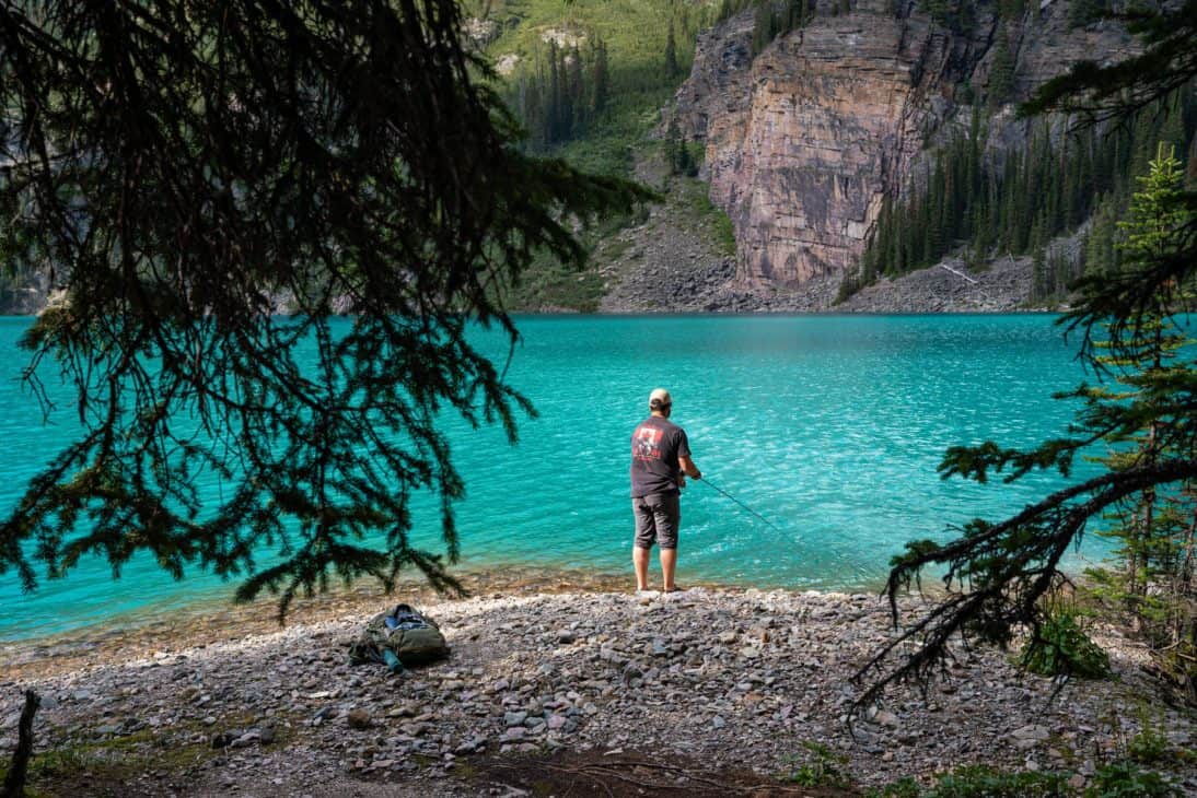 Photo of a man fishing on the shore of a deep turquoise blue lake with tree branches in the foreground. Fishing is one of the top activities in Lake O'Hara. 