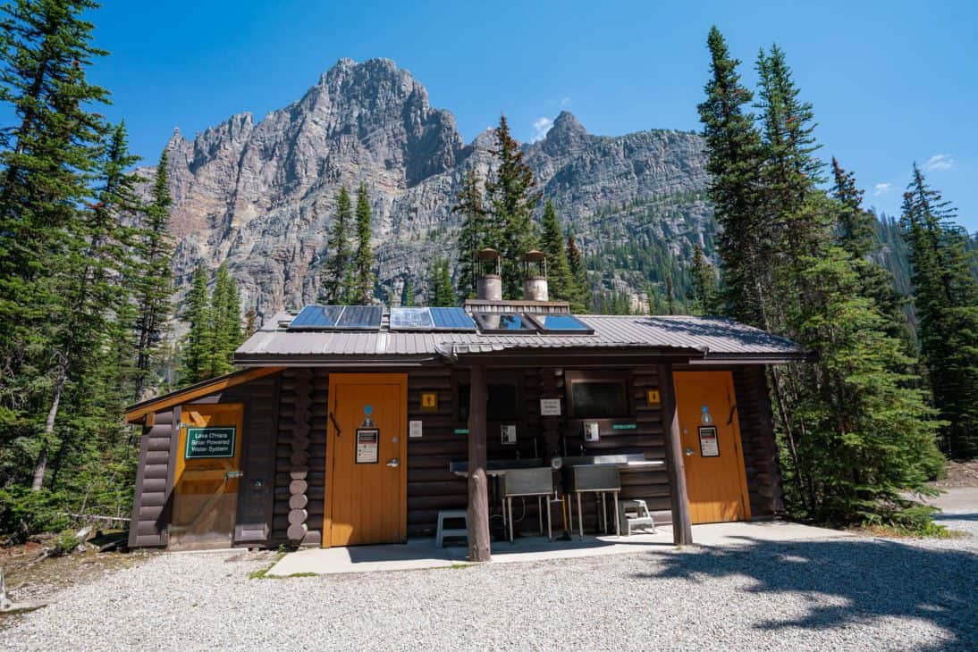 Photo of a log building with two sinks in front and solar panels on roof. Fir trees are either side of the log building with a rocky tall mountain in the background. The bathrooms at Lake O'Hara are some of the best I've seen in the backcountry.