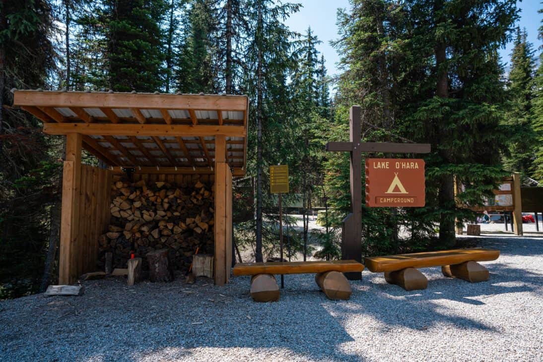 Photo of a shelter filled with chopped firewood next to two wooden benches with a sign that says "Lake O'Hara Campground"