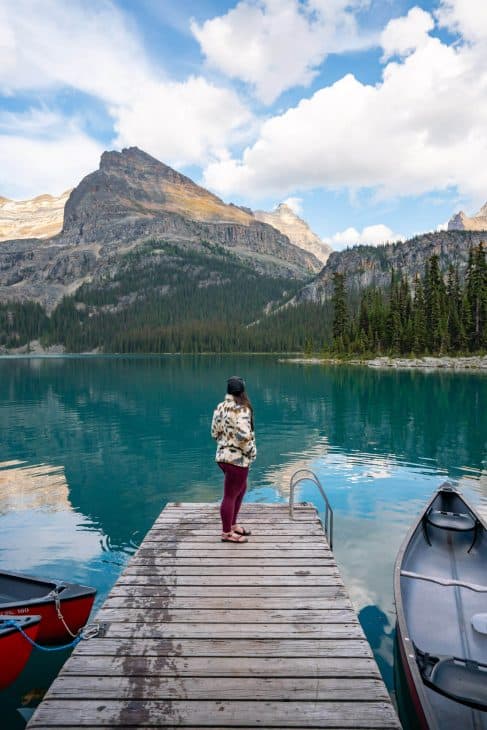 Photo of a boat dock with a woman standing at the end looking out towards the turquoise blue lake and looking and the mountain peak in the background.