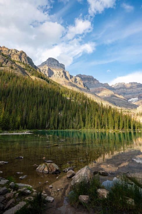 Photo of a clear green lake surrounded by bright green trees and rocky tall mountain peaks.
