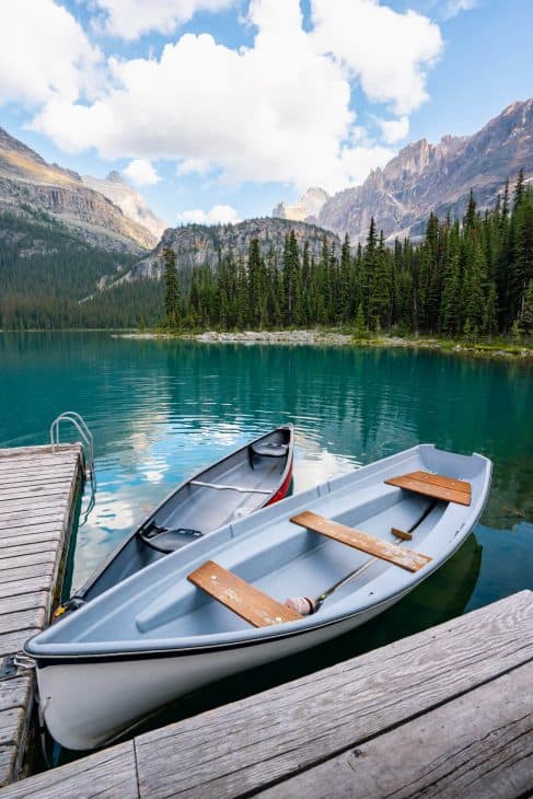Photo of a white canoe and a red canoe docked on a turquoise lake with trees and mountains in the background. 