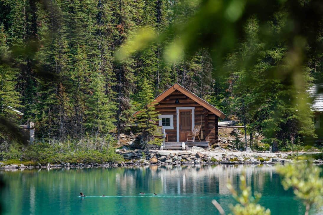 Photo of a small log cabin with chairs outside on the shores of a turquoise blue lake surrounded by green trees. In the foreground is a family of ducks swimming in the lake.