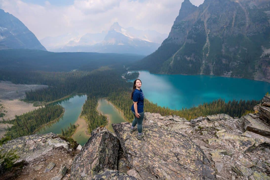 Photo of a woman standing on. a rocky lookout and smiling with her eyes closed. Below are two blue lakes surrounded by trees and mountains in the background.
