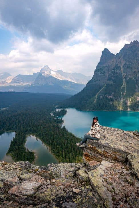Photo of a woman sitting on a rock ledge overlooking 2 turquoise blue lakes surrounded by fir trees and mountains in the background. The Opabin Plateau is the most iconic view of Lake O'Hara.