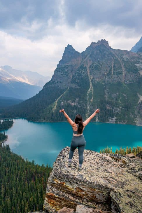 Woman standing on a rock ledge with her arms up looking out towards an alpine turquoise lake below and a mountains landscape. 