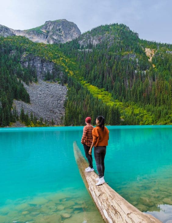 photo of a couple standing on a log suspended in the water of a turquoise lake with mountain peaks in the background.