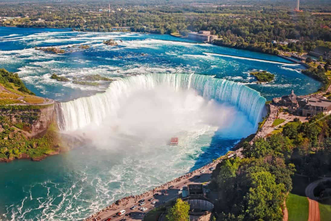 Photo from above Niagara Falls, a wide horseshoe shaped waterfall with a boat at the base of the falls sailing into the mist.