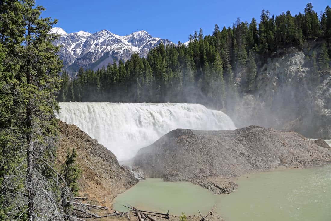 Long waterfall surrounded by fir trees and snow capped mountain peaks in the distance. Wapta Falls is one of the best hikes and things to do in Yoho National Park. 