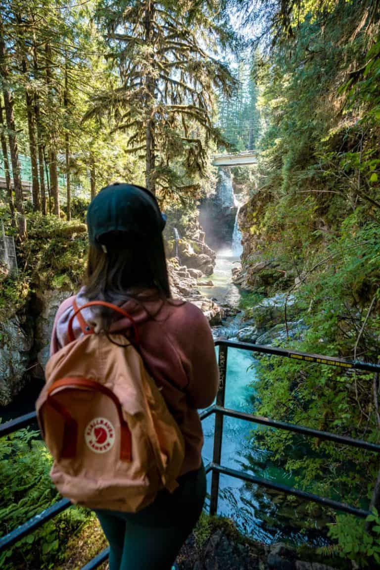 A woman wearing a backpack and a cap standing at a lookout platform surrounded by forests and admiring the view of a small waterfall in the distance.