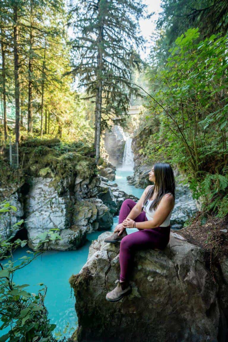 A woman sitting on a rock overlooking a bright blue river with a waterfall in the background in a lush rainforest. Mamquam Falls is one of the easiest hiking trails in Squamish.