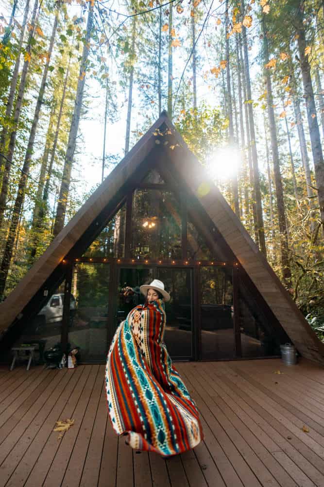 Photo of a woman with a fedora hat on and wrapped in a colourful blanket standing in front of an A Frame cabin. The Sky Haus A Frame cabin in Skykomish Washington is a great place to base yourself when visiting the Scenic Hot Springs