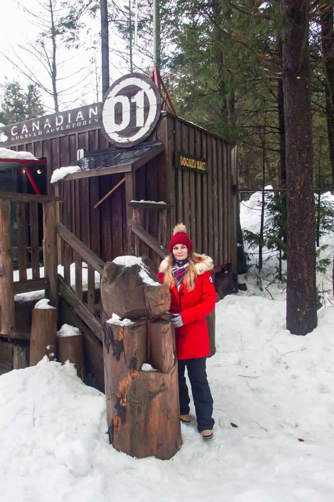 Photo of a woman wearing a bright red coat standing in front of a brown rustic cabin with snow covering the ground.
