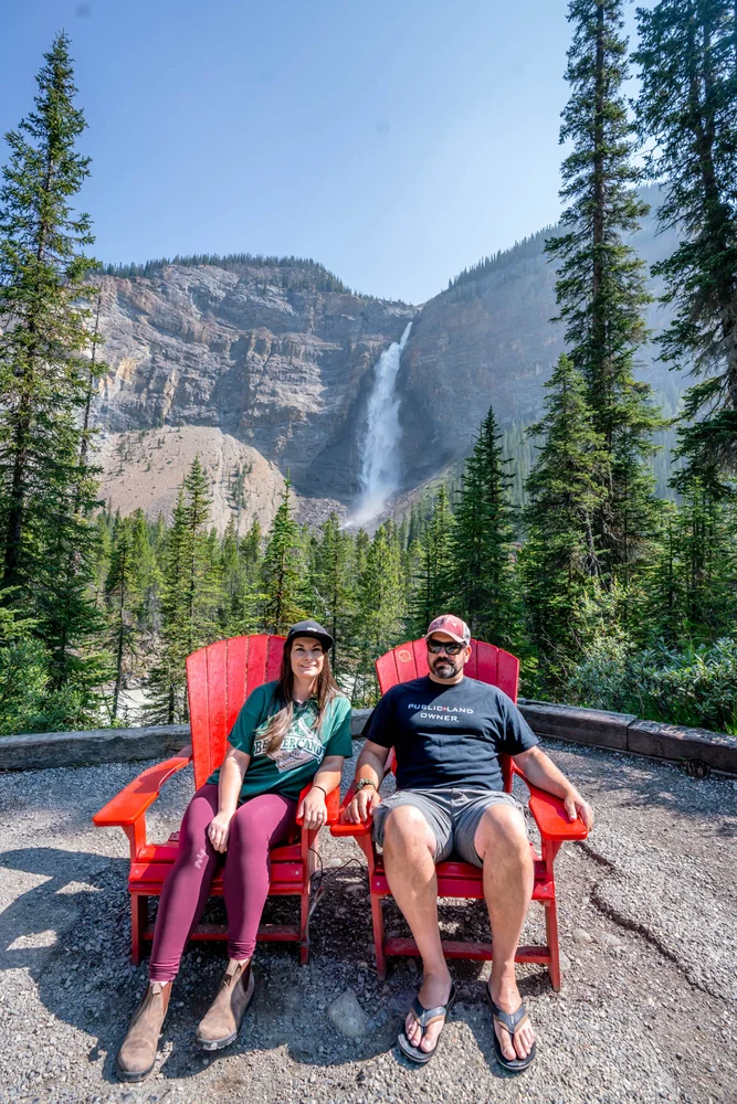 Rachael and Matt sitting in the red Adirondack chairs at Takakkaw Falls in Yoho National Park. 
