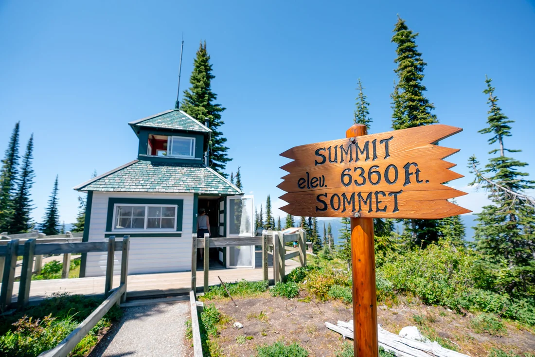 Photo of a white fire lookout with a green roof, in the foreground is a sign stating "Summit elev. 6360 ft. sommet"